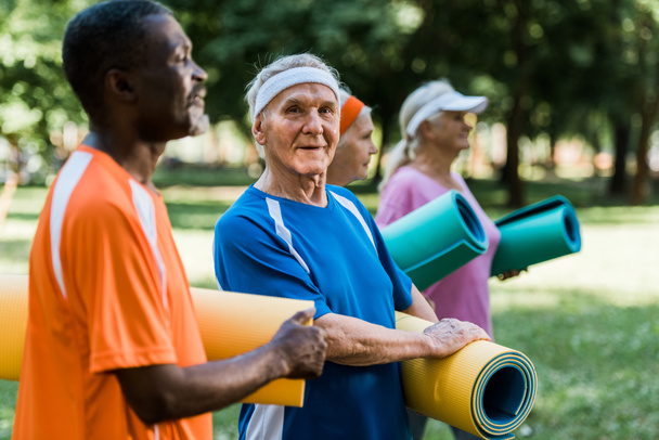 selective focus of happy multicultural senior pensioners standing with fitness mats in park  - Foto, immagini