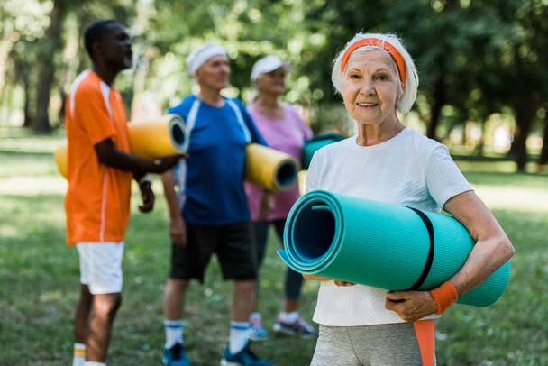 selective focus of happy retired woman with fitness mat smiling near multicultural pensioners  - Valokuva, kuva