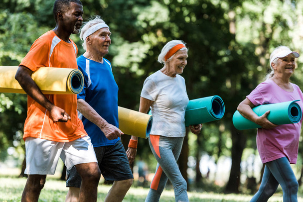 selective focus of positive retired and multiethnic pensioners holding fitness mats while walking in park  - Photo, Image