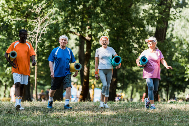 pensionistas multiculturales positivos y jubilados con colchonetas de fitness y caminando por el parque
  - Foto, Imagen