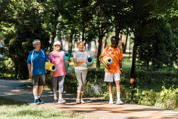 cheerful retired and multicultural pensioners holding fitness mats and walking in park  - Foto, Imagem