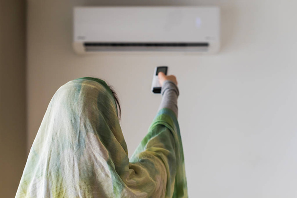 Muslim woman in a scarf  holding remote controller from air conditioner inside the room. - Foto, Imagem