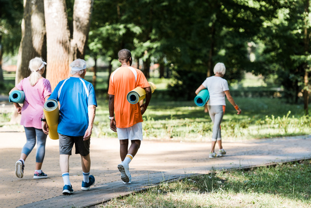 vue arrière des retraités heureux âgés et multiculturels tenant des tapis de fitness et marchant sur l'herbe
  - Photo, image