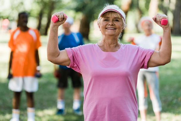 selective focus of happy retired woman in cap holding dumbbells while exercising near pensioners  - Photo, Image