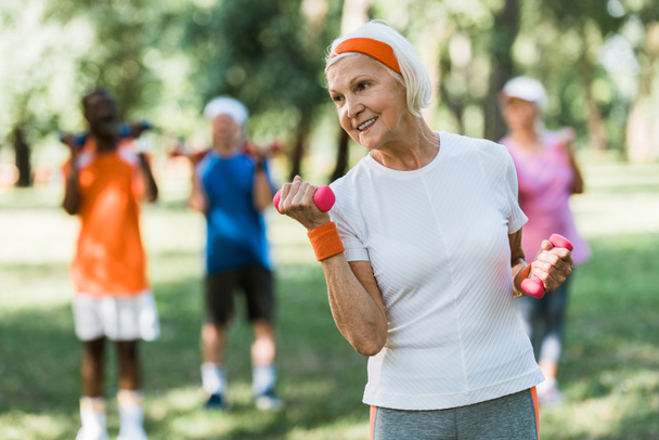 enfoque selectivo de la alegre mujer mayor sosteniendo pesas mientras hace ejercicio cerca de los pensionistas
  - Foto, Imagen