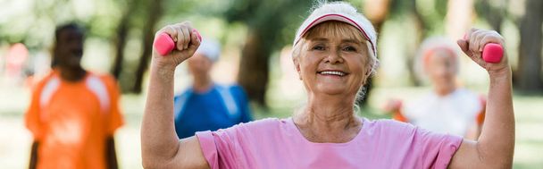 tiro panorâmico de mulher idosa alegre em boné segurando halteres durante o exercício perto de aposentados
  - Foto, Imagem