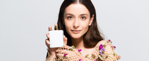 beautiful smiling woman in mesh clothing with purple flowers holding perfume bottle with copy space and floral scent isolated on grey - Fotografie, Obrázek