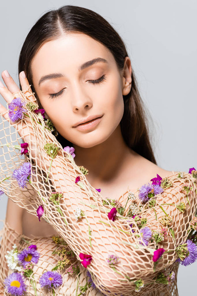 όμορφη γυναίκα με κλειστά μάτια στο δίχτυ μπεζ ρούχα με μωβ λουλούδια που παρουσιάζουν απομονωμένη σε γκρι - Φωτογραφία, εικόνα