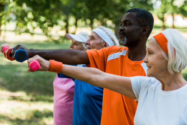 selective focus of african american man exercising with dumbbells with retired people in park  - Photo, Image