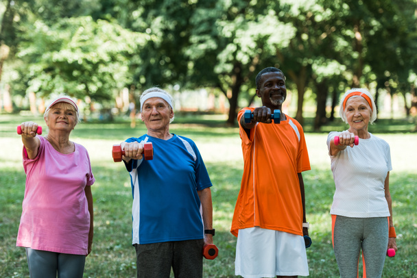 улыбающиеся мультикультурные пенсионеры с гантелями в парке
  - Фото, изображение