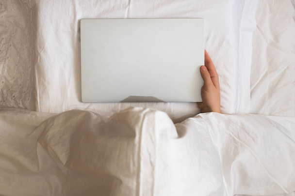 Girl lying with a laptop in bed covered by blanket. Woman can't sleep and have to work late at night. Top view - Foto, imagen