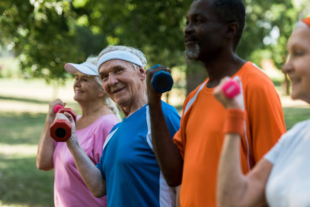 selective focus of happy senior man looking at camera while exercising with dumbbell near multicultural pensioners  - Foto, Imagen