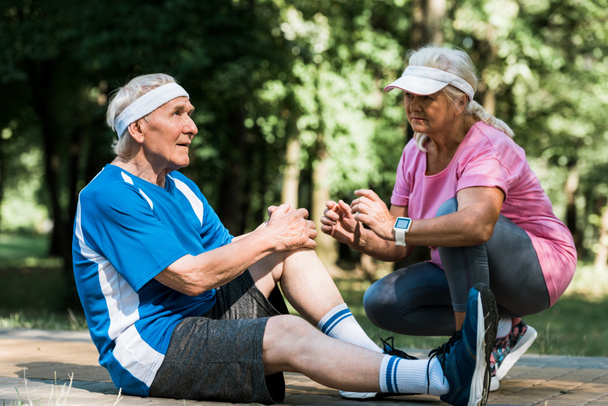 retired man touching knee while sitting on walkway near senior woman in cap  - Φωτογραφία, εικόνα