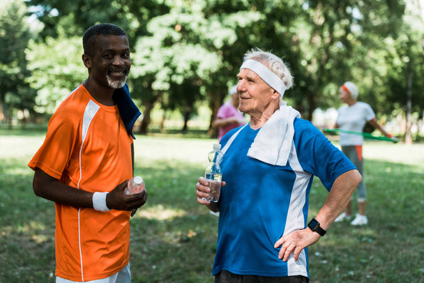 selective focus of sportive multicultural senior men holding bottles with water - Фото, изображение