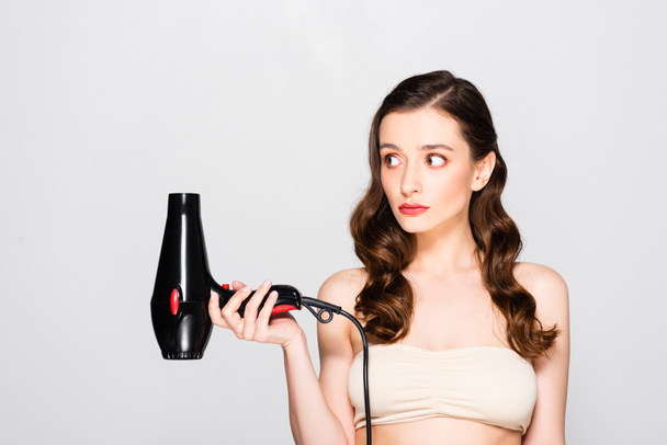 displeased beautiful brunette woman with curls and makeup holding hairdryer isolated on grey - Foto, imagen