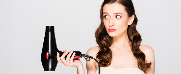 displeased beautiful brunette woman with curls and makeup holding hairdryer isolated on grey, panoramic shot - Photo, Image