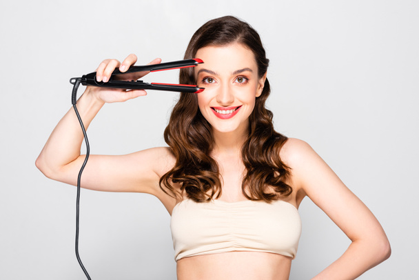 smiling beautiful brunette woman with curls and makeup holding straightening flat iron isolated on grey - Photo, Image