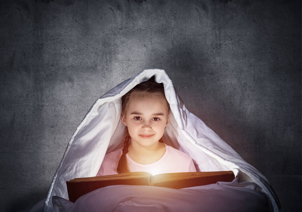 Happy little girl reading book in bed before going to sleep. Beautiful child with flashlight hiding under blanket. Portrait of smiling girl in pajamas on background of wall. Child reading fairy tales. - Foto, Imagen