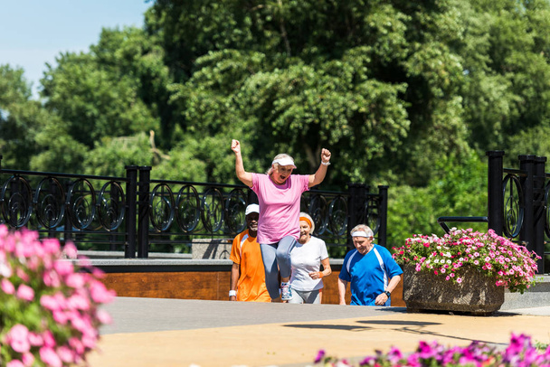 enfoque selectivo de la feliz mujer jubilada celebrando el triunfo cerca de los pensionistas multiculturales en el parque
  - Foto, Imagen