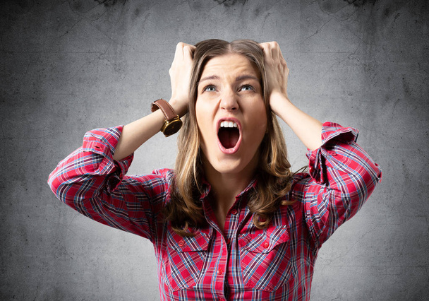 Stressful young woman screaming with panic and keeps hands on head. Emotional girl has irritated facial expression. Portrait of woman wears red checkered shirt on grey background. Panic attack - Фото, изображение