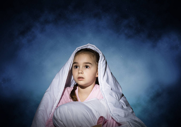 Scared girl with pillow hiding under blanket. Startled kid lying in his bed at home. Little girl afraid of dark. Covered child not sleep at night. Girl in pajamas on background of deep blue sky. - Fotoğraf, Görsel
