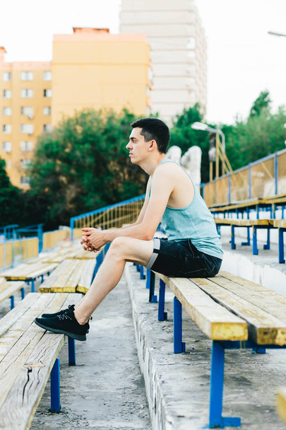 Bir bankta oturan ve ayakkabı bağcıklarını temiz havaya bağlayan genç bir adam. - Fotoğraf, Görsel