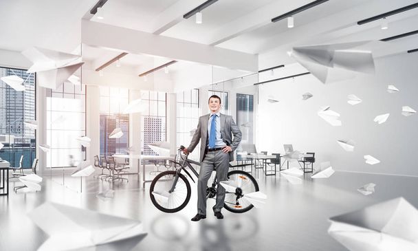 Smiling man in business suit standing with bike at comfortable conference room. Happy businessman with bicycle at modern office interior with big windows. Loft coworking space with flying paper planes - Фото, изображение