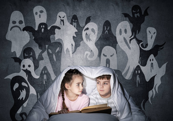 Cute girl and boy reading kids book in bed. Children with flashlight lying under blanket together. Kids in pajamas and funny ghosts silhouettes back on grey wall. Children reading magic fairy tales - Foto, Imagen
