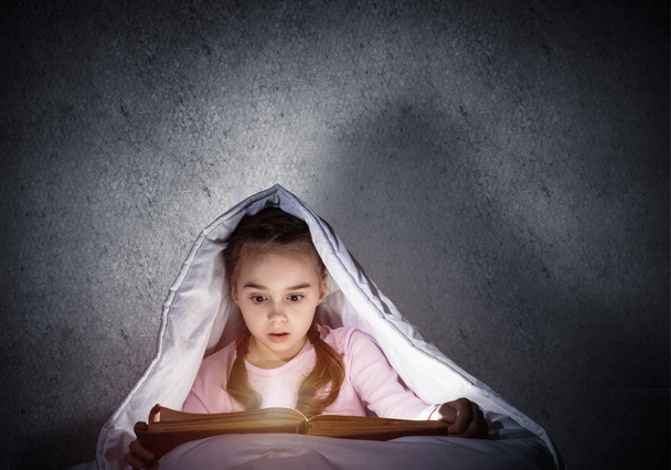 Engrossed little girl reading book in bed before going to sleep. Pretty kid with flashlight hiding under blanket. Beautiful girl in pajamas on background of grey wall. Child reading fairy tales. - Fotoğraf, Görsel