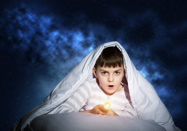 Surprised child with flashlight hiding under blanket. Amazed kid lying in bed at home. Fear of the dark. Little boy can not sleep at night. Portrait of boy in pajamas on background of deep starry sky - Photo, image