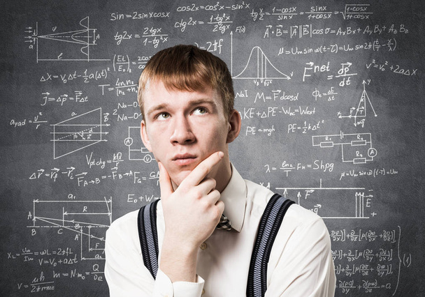 Ο κοκκινομάλλης μαθητής κοιτάζει προς τα πάνω και προσπαθεί να θυμηθεί κάτι. Ένα μπερδεμένο αγόρι που κάνει μαθηματικούς υπολογισμούς. Νέος επιστήμονας στο παρασκήνιο του chalkboard με μαθηματικούς τύπους. - Φωτογραφία, εικόνα