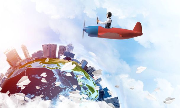 Man in aviator hat with goggles driving propeller plane. Earth globe with high modern buildings. Funny man having fun in small airplane. Blue cloudy sky with flying hot air balloons and paper planes - Photo, Image