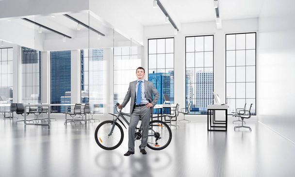 Smiling man in business suit standing with bike at design studio with computer workstations. Happy businessman with bicycle at modern office interior with panoramic windows, desks and chairs - Foto, Imagen