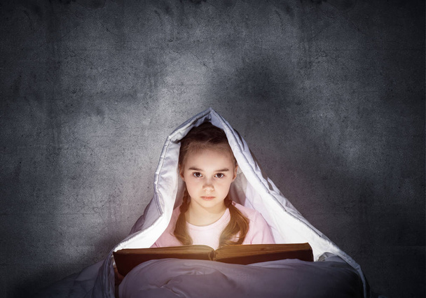 Una niña leyendo un libro en la cama antes de irse a dormir. Niño bonito con linterna escondida debajo de la manta. El chico no puede dormir por la noche. Retrato de hermosa chica en pijama sobre fondo de pared gris
. - Foto, Imagen