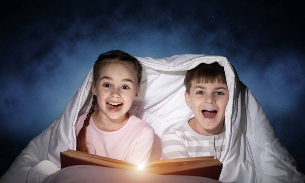 Boldog gyerekek zseblámpával bujkál alatt takaró együtt. Mosolygós gyerekek olvasás tündérmesékben az ágyban. Kis barátok szórakoztunk leesti. Fiatal fiú és lány pizsama nem alszik éjjel. - Fotó, kép