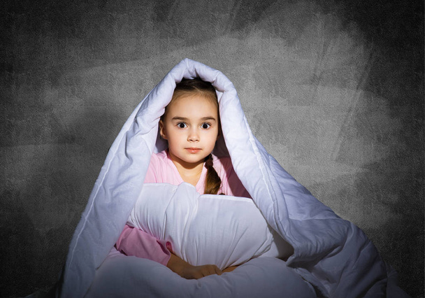 Frightened girl hiding under blanket. Scared kid lying in his bed at home. Fear of the dark. Beautiful little girl can not sleep at night. Portrait of child in pajamas on background of grey wall. - Valokuva, kuva