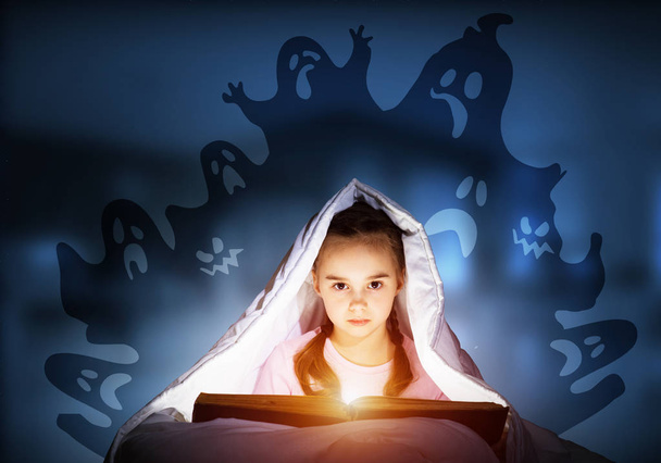 Scared kid with reading book under blanket. Afraid girl lying in bed at home. Child reading scary stories. Girl in pajamas and imaginary ghosts back on night sky. Bright light shining from open book. - Foto, Bild