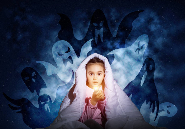 Scared girl with flashlight hiding under blanket from imaginary ghosts. Kid sitting in bed on night sky background. Covered child in pajamas not sleep at night. Mysterious phantoms in darkness. - Foto, imagen
