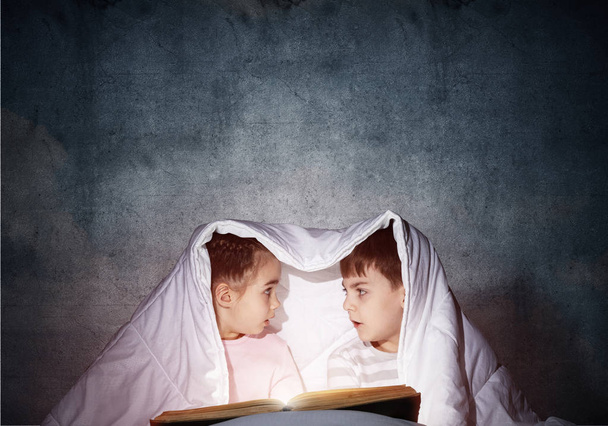 Surprised children reading scary stories in bed before going to sleep. Amazed girl and boy in pajamas hiding under blanket together. Covered kids with open book not sleep at night. Happy childhood - Foto, Imagen