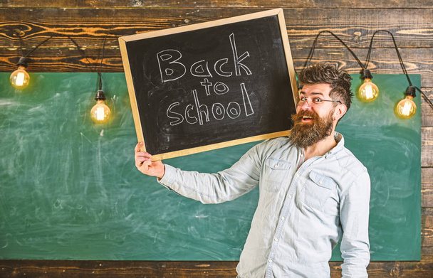 High school concept. Man with beard and mustache on cheerful face welcomes students, chalkboard on background. Teacher in eyeglasses holds blackboard with title back to school - Foto, imagen