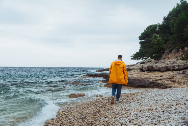 homem andando pela praia rochosa em capa de chuva amarela. tempo ventoso nublado
 - Foto, Imagem