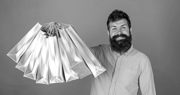 Man with beard and mustache holds shopping bags, red background. Hipster on happy face is shopping addicted or shopaholic. Guy shopping on sales season with discounts. Shopping concept - Photo, Image