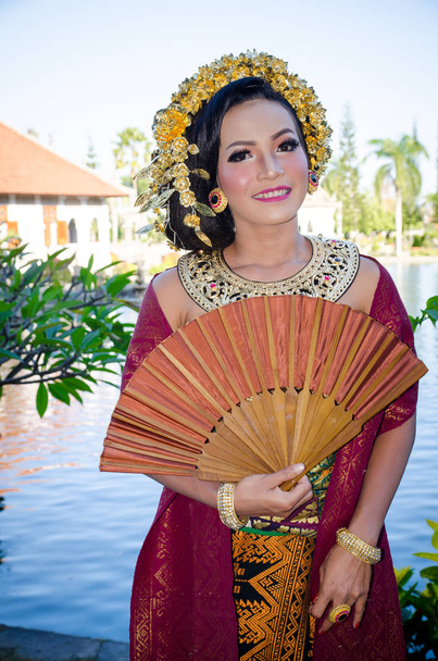 Красивая балийская девушка в традиционном платье в парке, портрет с ручной вентилятор
 - Фото, изображение