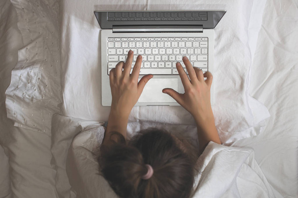 Girl lying with a laptop in bed covered by blanket. Woman can't sleep and have to work late at night. Top view - Photo, Image