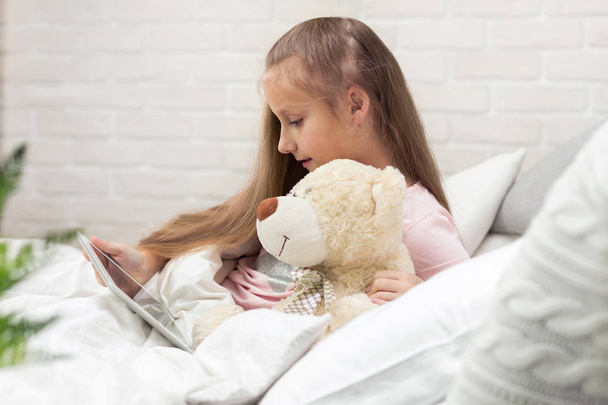 cute little girl lies in bed uses digital tablet. - Foto, afbeelding