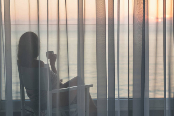 nackt schöne Frau in eine Decke gehüllt auf einem Stuhl sitzend mit einer Tasse Kaffee hinter den transparenten Vorhängen auf dem gläsernen Balkon frühmorgens bei Sonnenaufgang. Meerblick - Foto, Bild