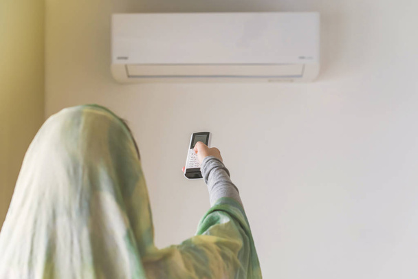 Muslim woman in a scarf  holding remote controller from air conditioner inside the room. - Valokuva, kuva
