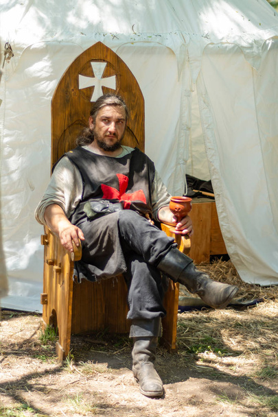ニス、セルビア - 6月15日。2019年:民族衣装を着た騎士は、テントの前で木製の玉座に座り、騎士の国際フェスティバルでワインを飲みます。クローズアップ、選択的フォーカス - 写真・画像