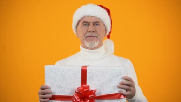 Positive male pensioner in santa claus hat shaking giftbox and smiling, surprise - Filmagem, Vídeo