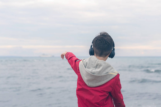 A boy in a red jacket and headphones looks at the sea waves in stormy rainy weather - Foto, Imagem